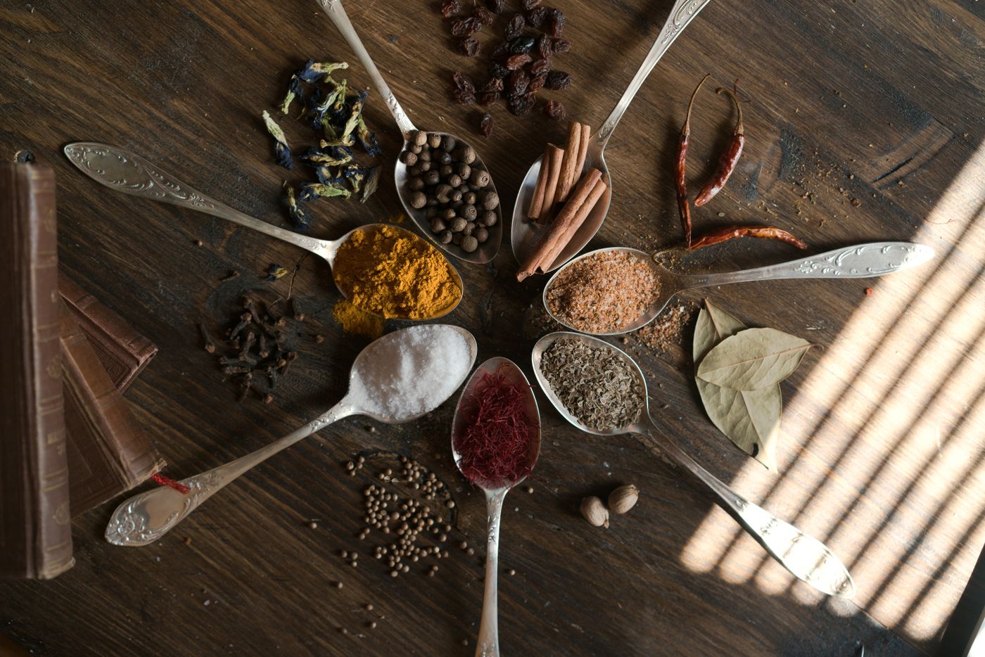 Silver spoons of turmeric, saffron, cloves, cinnamon, peppercorns and bay laid out on dark wood in slatted window light.
